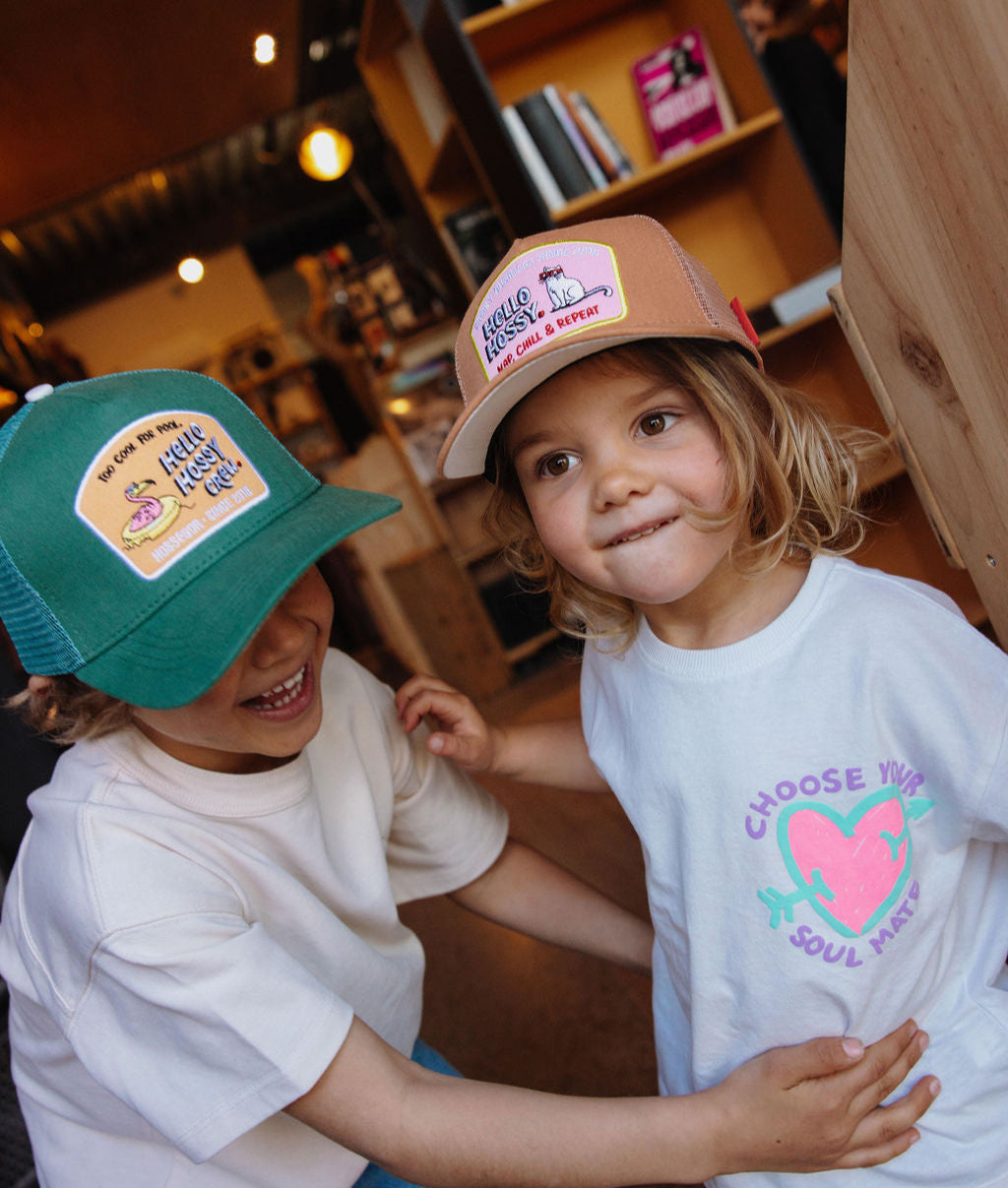 Boné infantil castanho com pala reta e patch Hello Hossy, usado por uma criança Hello Hossy