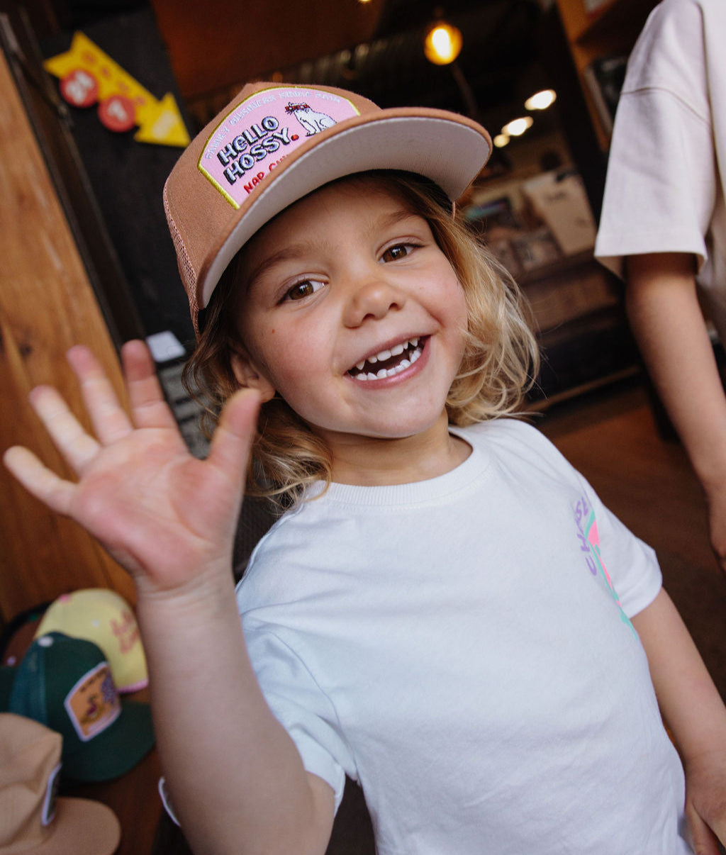 Boné infantil castanho com pala reta e patch Hello Hossy, usado por uma criança Hello Hossy