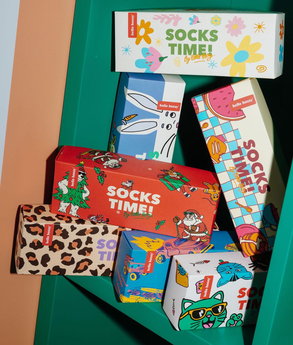 Caixas de presente "Socks Time!" empilhadas numa prateleira verde, embalagens coloridas e ilustradas: padrões de natal, flores, leopard, mascotes, carros, gatos e comida – Hello Hossy