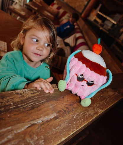 Peluche em forma de bolo com óculos de sol Cherry Cake, pousado numa rampa com uma criança a olhar para ele - brinquedo original - Hello Hossy