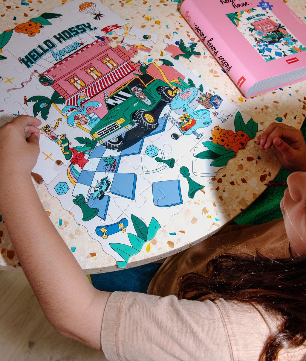 Menina a montar o Puzzle colorido de 48 peças "Hello Hossy House" numa mesa – vista de cima – Hello Hossy