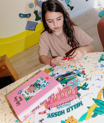 Menina a montar o Puzzle ilustrado "Hello Hossy House" numa mesa, com a caixa ao lado – fotografia lifestyle – Hello Hossy