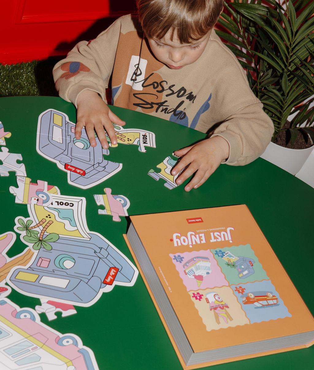 Rapaz com um dos quatro puzzles "Just Enjoy" parcialmente montado, a caixa e a ilustração-modelo, sobre uma mesa à sua frente – foto lifestyle – Hello Hossy