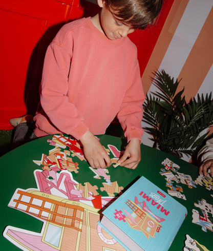 Criança a brincar com o Puzzle de 28 peças "Miami Plage" numa mesa, com a caixa ao lado – ambiente divertido – Hello Hossy