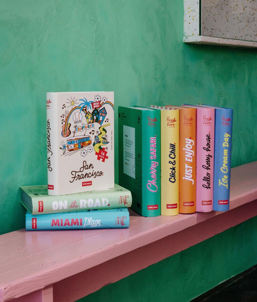 Coleção de puzzles Hello Hossy pousados num banco rosa – design com efeito de livro – fundo de parede colorido