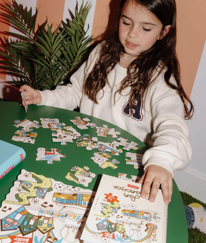 Menina a montar o Puzzle colorido de 16 peças "San Francisco" numa mesa – vista frontal – Hello Hossy